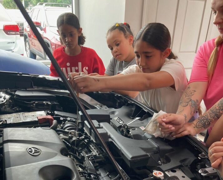 girl checking the oil of a vehicle as two girls and instructor look on